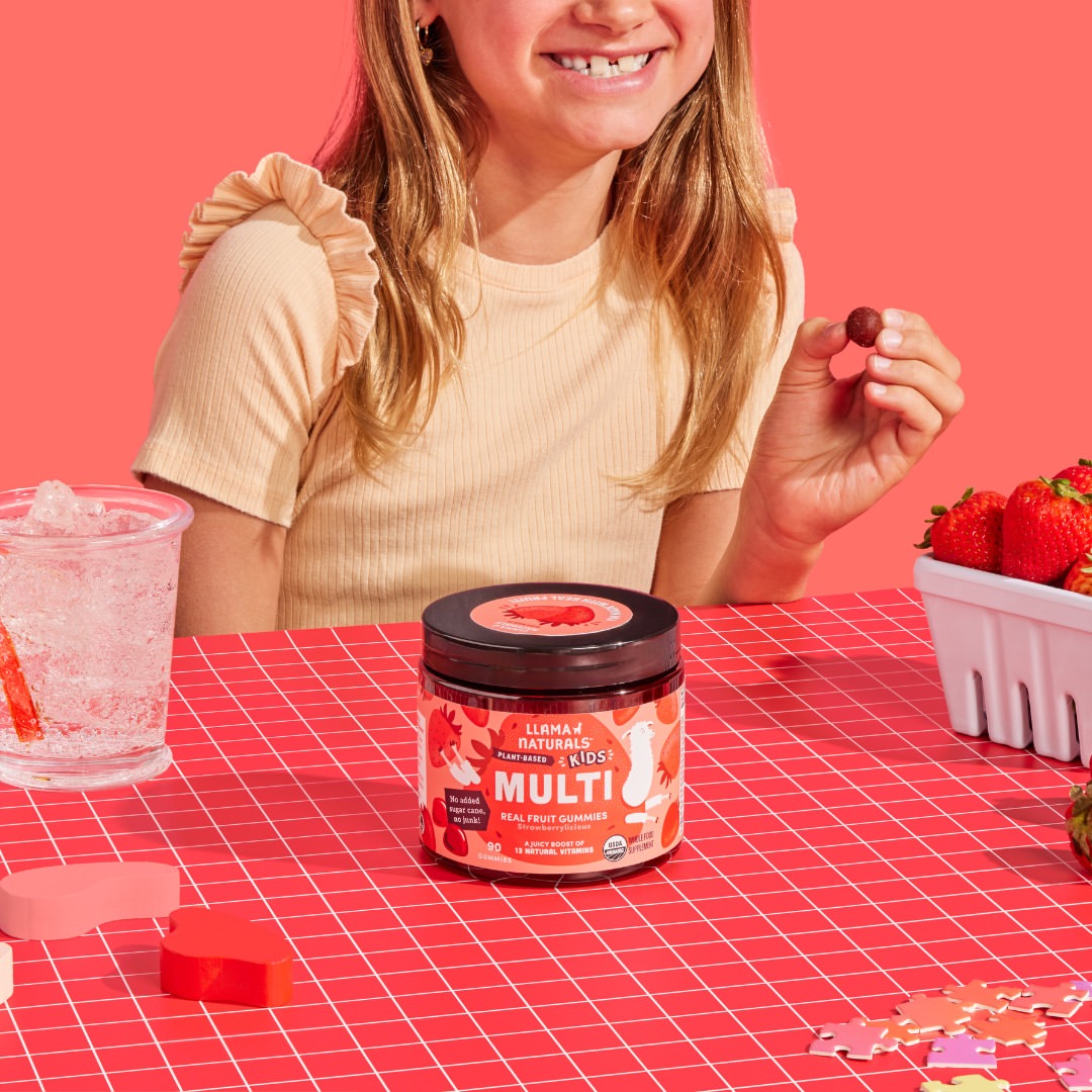 Smiling girl at a kitchen table reaching for a Llama Naturals gummy, jar and fresh strawberries nearby